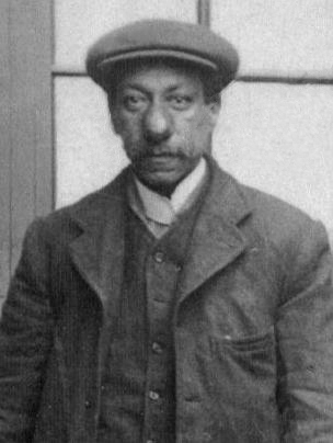 Head-and-shoulders photograph of a middle-aged man in a flat cap, collar and heavy jacket.