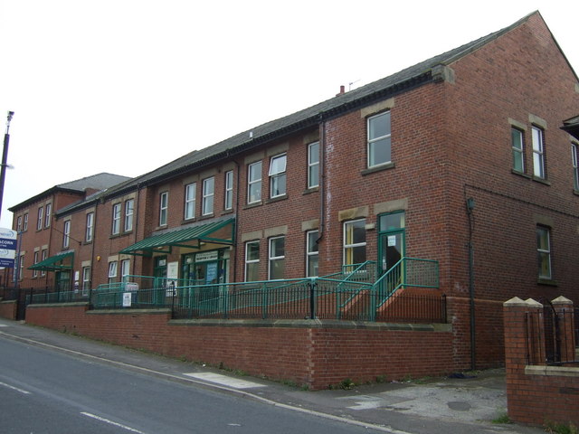 A long two-storey red-brick building with green railings and canopies, on a sloping street in Grimethorpe.