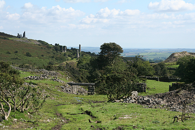 A grassy moorland slope with stone walls and the ruined chimneys and engine houses of a copper mine standing along the hillside.