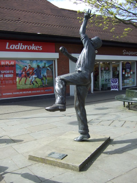 A bronze statue of a cricketer in his bowling stride on a stone plinth in a paved market place.