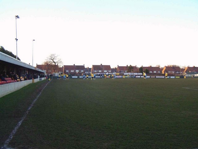 A non-league football ground on a winter afternoon: a covered stand along one touchline, advertising boards, houses behind, and players in yellow and blue on the pitch.