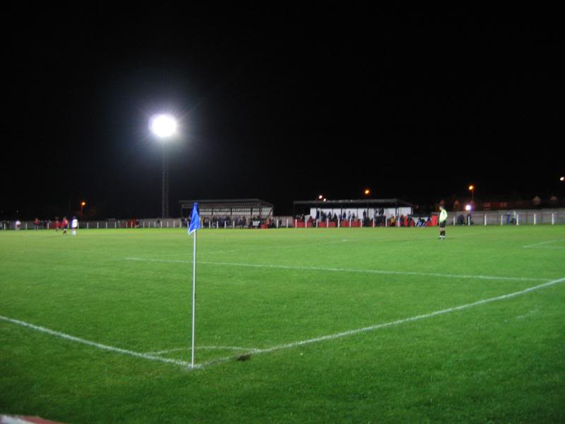 A floodlit football pitch at night with a corner flag in the foreground and a low covered stand and seated stand across the far touchline.