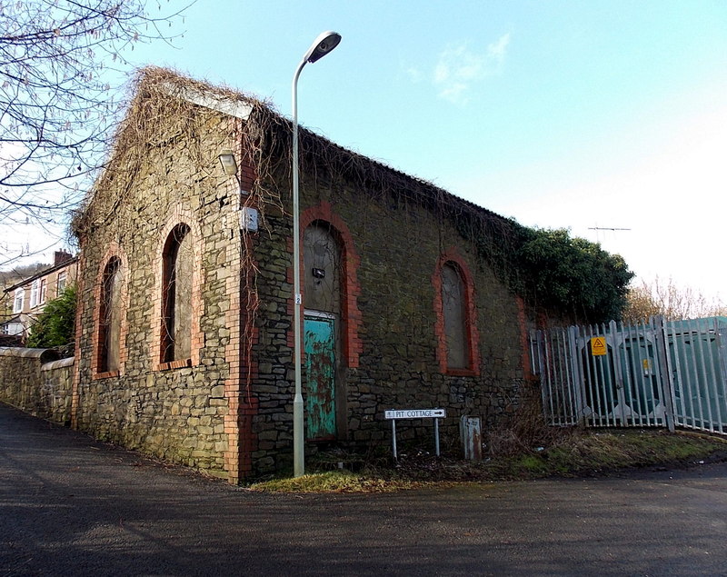 A derelict stone colliery building with arched, brick-edged windows and ivy on a street corner.