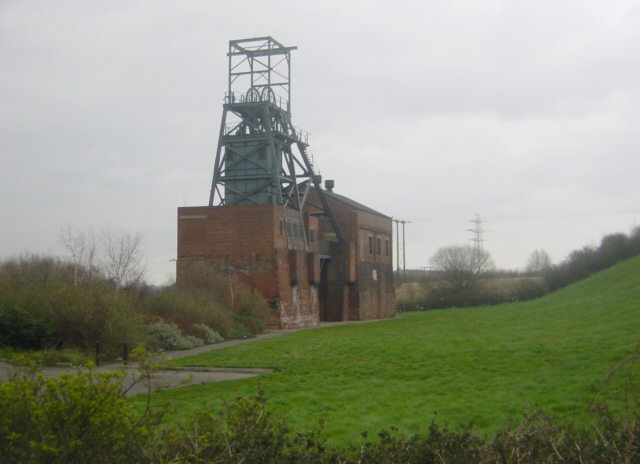 A steel headframe above a brick engine house beside a grassy bank under a grey sky.