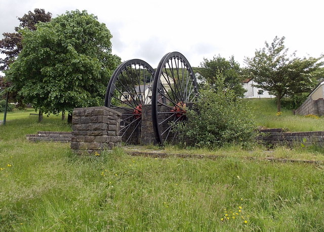 Two black colliery winding wheels set on a stone plinth in grass, with trees and houses behind.
