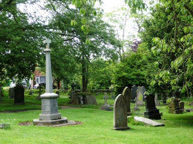 A tall stone cross on an inscribed pedestal among gravestones in a tree-lined churchyard.
