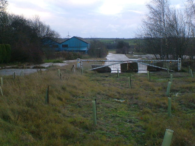 Rough grassland with young trees in guards, a gated track and a blue-roofed workshop beyond.