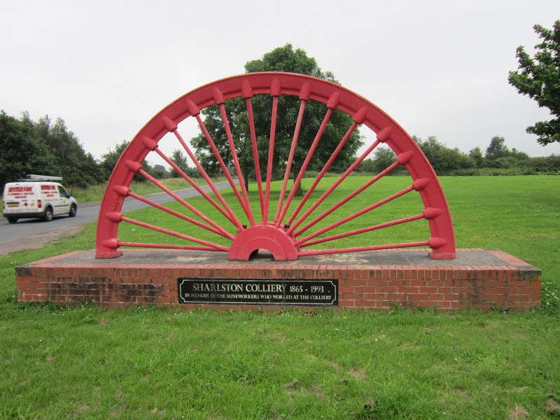 A red half pit-wheel memorial on a brick plinth lettered Sharlston Colliery 1865-1993, on a grass verge.