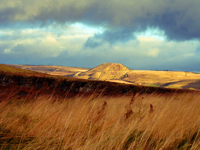A conical colliery spoil tip on open moorland under a stormy sky, seen across rough grass.
