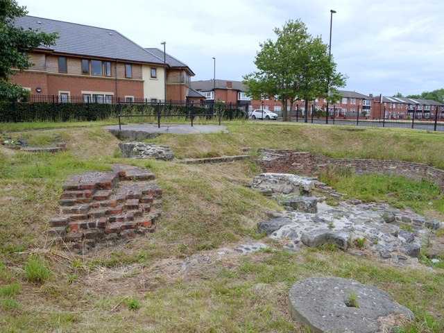 Excavated brick footings and stone remains of a colliery shaft site in grass, with houses beyond.