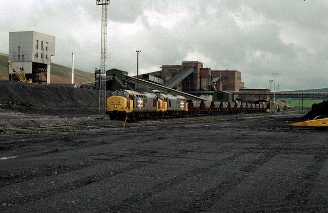Abernant Colliery in the late 1980s: a concrete headframe tower and the brick washery buildings behind a pair of diesel locomotives hauling loaded coal wagons across a black coal yard.