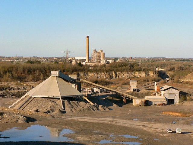A working limestone quarry: a long covered conveyor and stockpile in the quarry floor, the bench face behind, and the cement works with its tall chimney on the skyline.