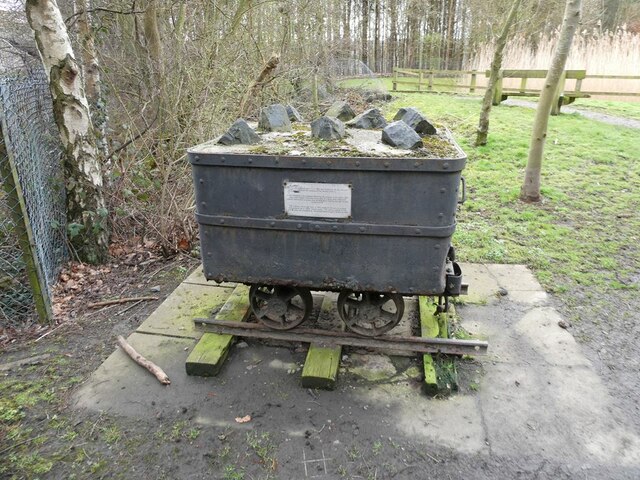 Riveted iron pit tub with a small plaque, topped with lumps of stone and set on a short length of rail beside a wooded path