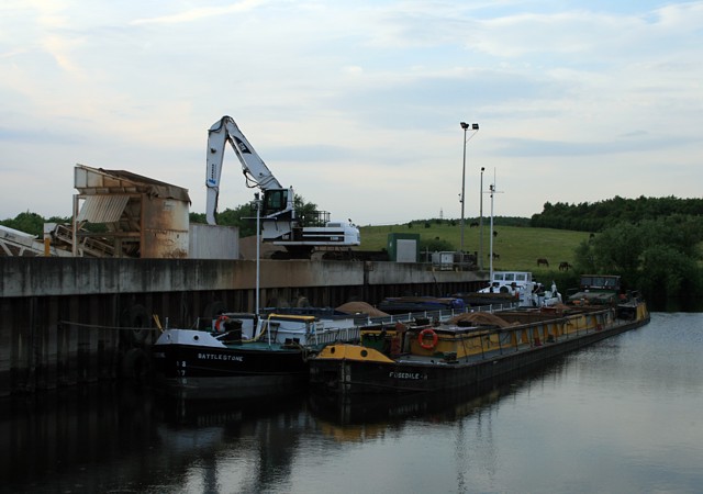 Two laden barges moored at a concrete river wharf with a crane and hopper, green hills beyond, at evening, 2008
