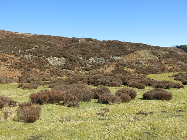 Heather-covered moorland with pale spoil heaps and broken ground along a valley side under a clear blue sky.