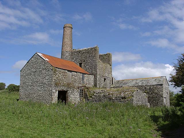 The roofless granite shell of a rotative beam winding engine house at Belowda, its tapered chimney standing beside it above rough ground