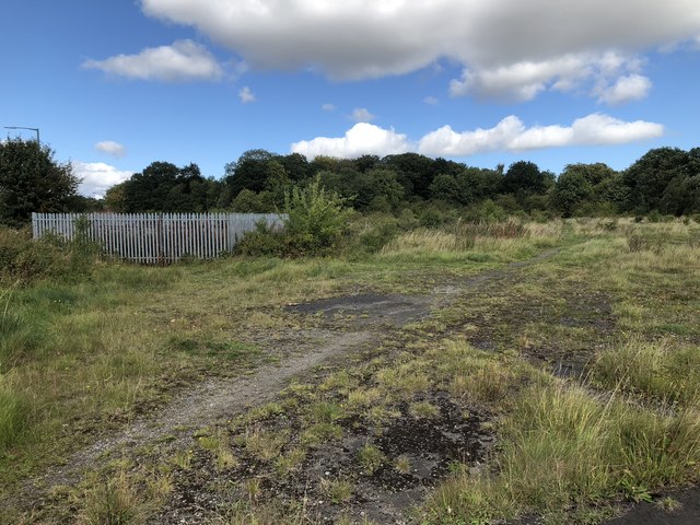 Rough grass and patches of old tarmac on waste ground, with a tall steel palisade fence enclosing part of it and trees along the horizon.