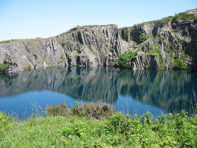 Sheer grey slate cliffs are reflected in a still, deep-blue flooded quarry pit under a clear sky.