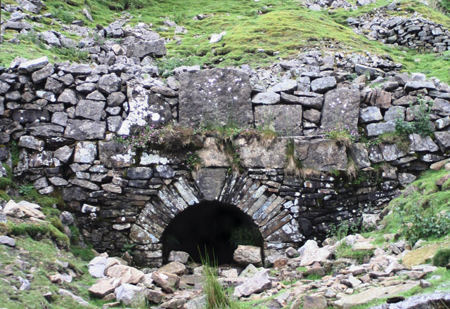An arched stone portal set into a revetment wall on a stony hillside, the mouth of the mine level, its floor choked with rubble.