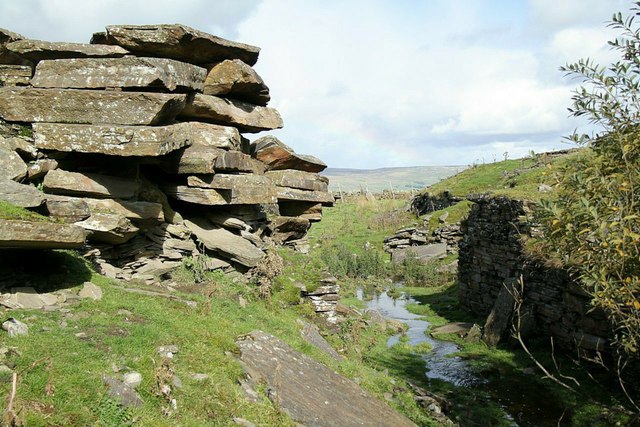 Thick slabs of pale sandstone stacked in ragged layers at the mouth of a level, with a small beck running past ruined drystone walling and open moor rising beyond.