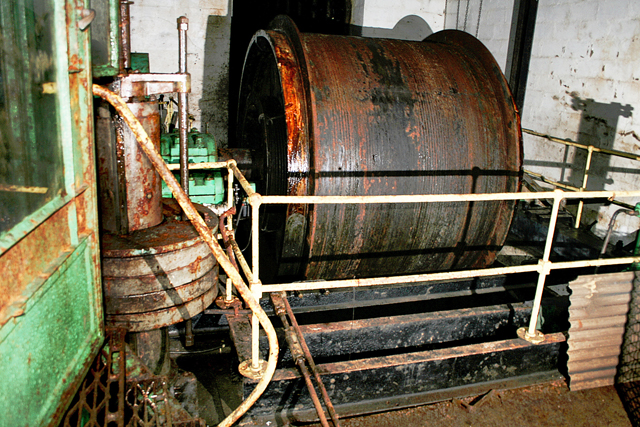 A large rusted winding drum and its green-painted machinery behind white safety railings in a whitewashed underground chamber.