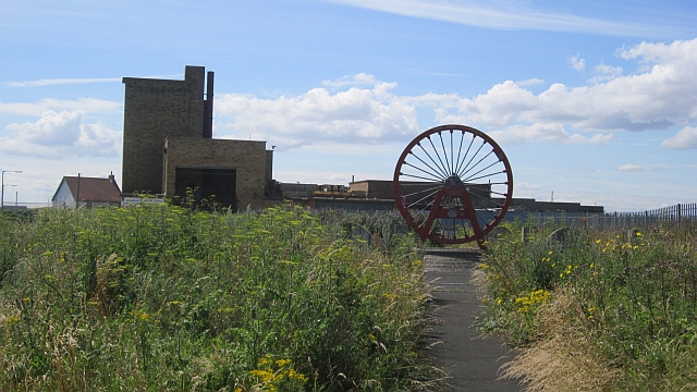 A red-painted colliery winding wheel standing as a monument at the end of a path through rough grass, with surviving pithead buildings behind.