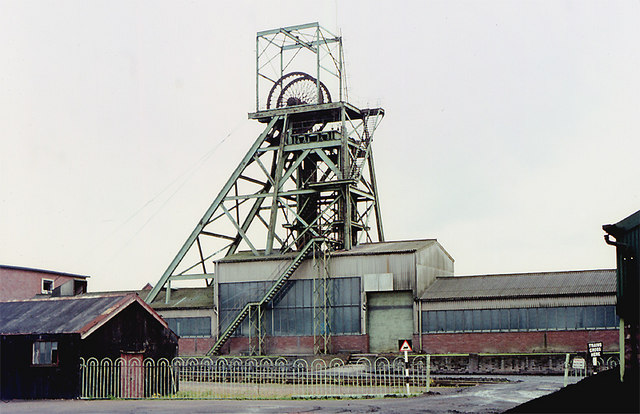 A steel pithead headframe above corrugated sheds and brick buildings behind a green fence.