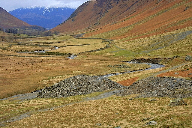 Grey spoil heaps from the lead mine on the valley floor beside a stream, with russet fellsides rising either side and a snow-capped fell beyond.