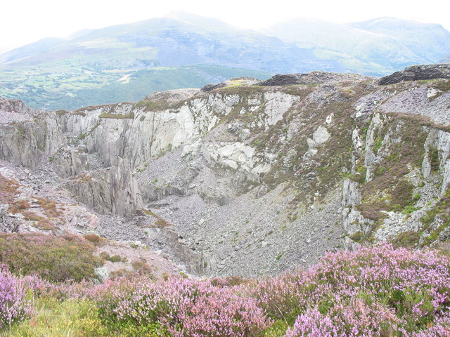 A deep rocky slate pit with pale grey walls and waste slopes, purple heather in front and mountains beyond.