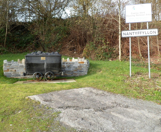 A restored iron coal dram mounted on a short length of rail beside a roadside verge, with a plaque set in front of it and hillside behind.