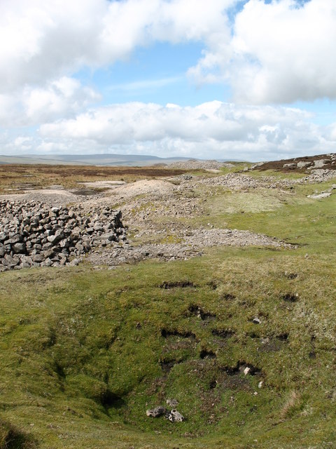 A grassy hollow of a collapsed shaft in front of stone walling and spoil tips on open moorland.