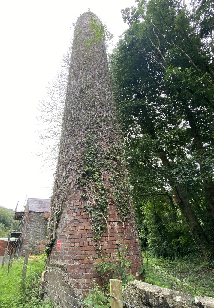A tall, tapering brick chimney covered in ivy, beside trees and a stone building.
