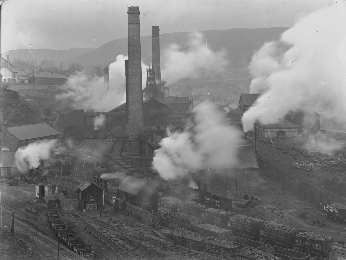 A historic photograph of Cymmer Colliery at Porth, showing the pithead buildings and winding gear in the valley.