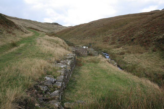 A grassy hollow beside a moorland stream with a long stone revetment wall and a ruined structure at its end, the valley rising on both sides.