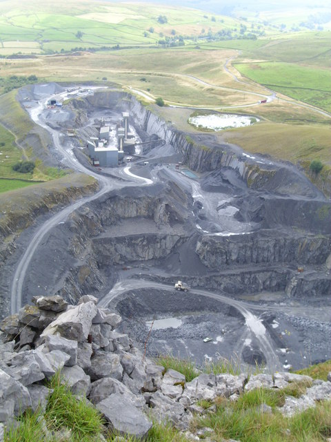Looking down into the working face of Dry Rigg Quarry from the limestone edge above, grey benched rock and haul roads with the dale beyond.