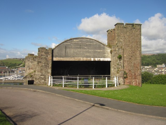 A stone fan house with a broad arched opening and a crenellated tower at each end, standing on grass above the harbour.