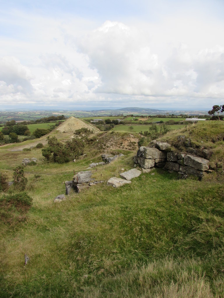 Grassy hollows and tumbled granite footings on a hillside, with a pale spoil heap and wide farmland beyond.