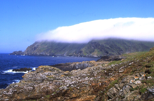 A low rocky headland with reddish-stained rock marking old mine workings, a cloud-capped hill beyond.