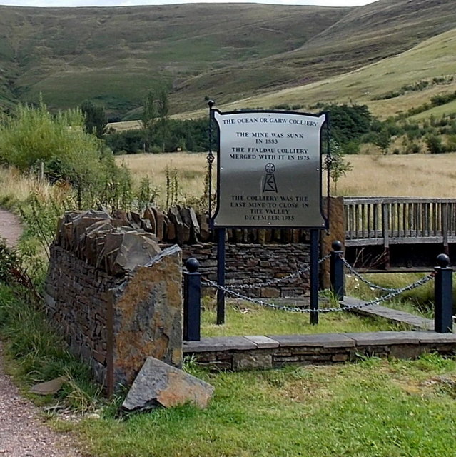 A framed memorial plaque on a stone plinth beside a path, recording the colliery's sinking and closure, with a valley of bare hillsides behind.