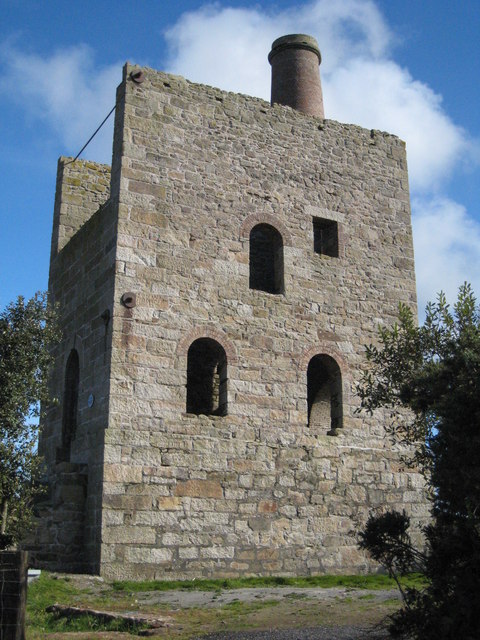 A tall granite engine house with arched window openings and a round chimney rising behind, under blue sky.