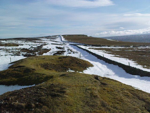 A snow-covered spoil heap and disused shaft at Wellington Shaft, on Fremington Edge Top, marking the western boundary of the Hurst Mining Field, with a stone wall and bridleway gate visible.