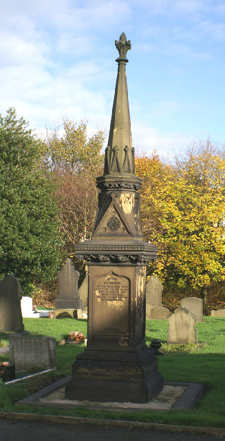 A tall dark stone memorial with a carved spire and inscribed panel standing among gravestones in a cemetery, with autumn trees behind, 2005