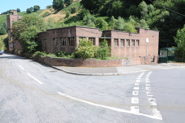 A long, flat-roofed brick and concrete bath house standing empty and boarded, with broken windows and grass growing up around it.