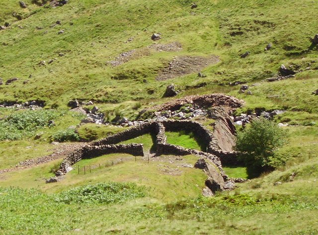 Drystone sheepfold walls built among rocky mining debris on a green fellside, with small spoil patches on the slope behind.