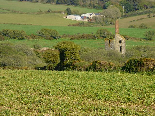 A green field in the foreground with the ruined engine house and chimney of Medlyn Moor among hedges and farmland beyond, 2019.