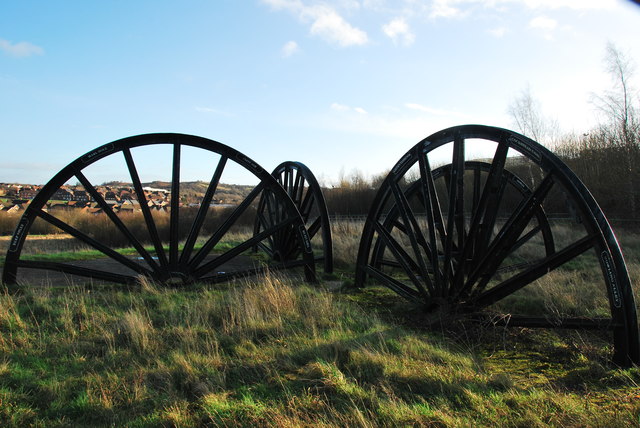 Three large black spoked pithead wheels set upright in rough grass on top of a landscaped mound, with houses in the distance, 2015