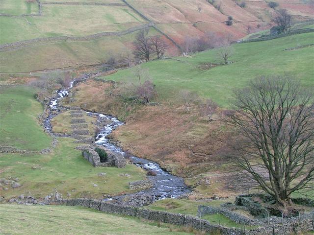 A stepped line of stone launder piers and a roofless wheelpit beside a rocky beck in a green valley bottom, below walled fields and bare trees.