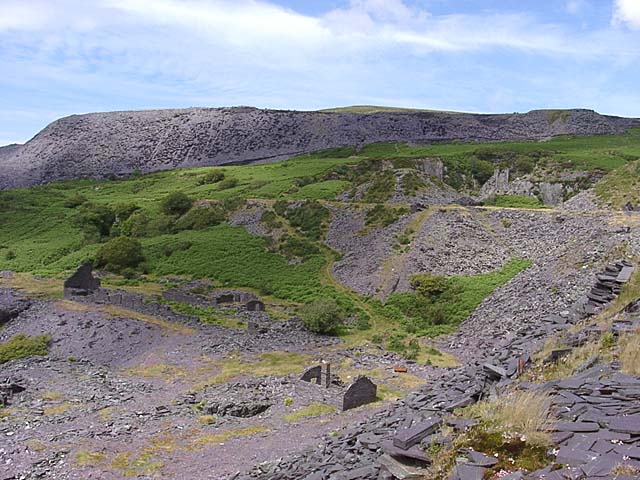 Roofless stone walls of a slate mill among grassed-over waste tips in a Welsh valley.