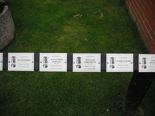 A row of small memorial plaques on a wooden rail set in grass, each with a miner's lamp and the name of a Polmaise miner.