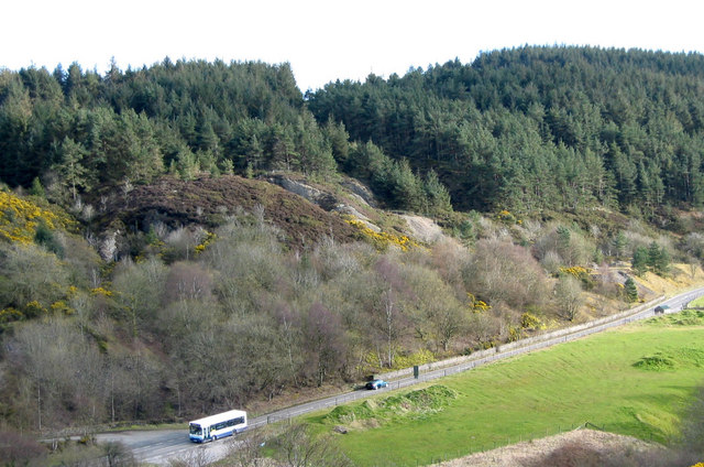 A wooded hillside scarred with pale spoil and old mine workings above the A488, with a bus on the road and a field below.
